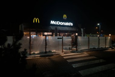 WARsaw, POLAND - 5 Ocak 2026: Geceleri aydınlatılan modern McDonald 's restoranının dış görünüşü. İkonik Altın Kemerler ve marka işareti binanın üzerinde parlıyor, buzlu cam bariyer ve yaya geçidi öngörülebiliyor.