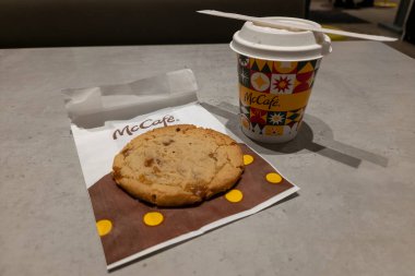 UYARI, POLAND - 5 HAZİRAN 2026: Bir McCafe kağıt kahve fincanının kapağı ve kağıt kamışla yakın plan perspektifi, markalı bir poşetin üzerinde bir kurabiyenin yanına yerleştirilmiş. Fast-food tatlı seti restoranın içindeki gri masada sergileniyor.