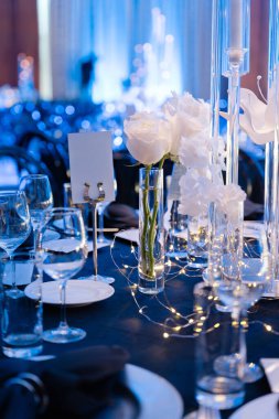 Beyaz güller, cam vazolar ve mavi ışıklı lüks salonda sıcak peri ışıkları olan zarif ziyafet masası düğün ya da gala için hazırlandı.