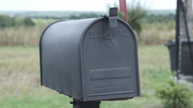 Kırsal alanda kırmızı bir bayrak taşıyan siyah posta kutusu, yeşil alanlar ve ağaçların bulanık arka planına karşı duruyor, geleneksel posta servisi ve iletişimi simgeliyor.