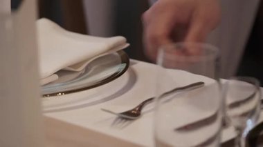 Masayı beyaz bir peçete, cam tabak ve gümüş çatal bıçakla hazırlayan eli kapat. Statik çekim.