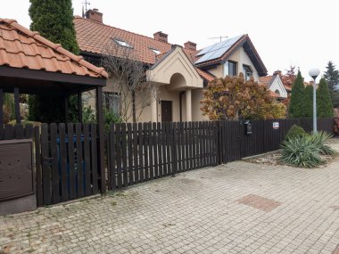 SAVAŞ, POLAND - 8 Kasım 2025: Kahverengi kiremit çatılı ve güneş panelleri olan, koyu ahşap bahçe çitleri ve kaldırımlı bir garaj yolu bulunan modern, tek aile evi