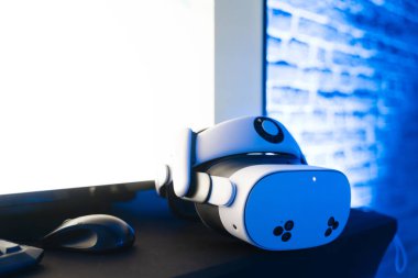 Beyaz sanal gerçeklik kulaklığı ve oyun pervazları mavi ışıklandırmalı karanlık bir masanın üzerinde.