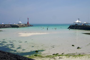 Bazalt resiflerine bakan Segial Beach 'teki korunaklı koy Gimnyeong' un altında soluk kum ve turkuaz suyla kaplanıyor. İkiz dalgalar, rıhtım çadırları ve yüzme alanını çevreleyen kırmızı bir fener..