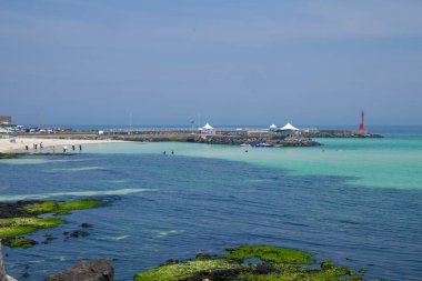 Gimnyeong 'un doğusundaki bazalt sahilinden Segial Beach, ikiz dalgalar arasında sığ bir lagünün içinde açılıyor. İnsanlar solgun kumların üzerinde yürürken kırmızı bir fener ve iskele çadırları koyları çerçeveliyor..