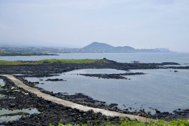 A straight concrete path crosses tidal pools and stone weirs on the Shinheung coast, with a cinder cone rising hazily beyond the shoreline.