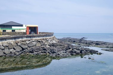 A renovated seaside house and garage sit above a curved seawall at Gimnyeong, facing shallow tidal pools and basalt reefs along the quiet East Sea coast.