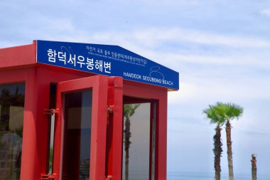 Kırmızı bisiklet sertifikası standının tepesindeki Hamdeok Seoubong Sahili 'nin iki dilli tabelasına yakından bakın. Palmiye üstleri ve burnun ötesinde görünen sakin deniz..