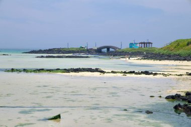 Shallow tidal flats stretch toward a small islet at Hamdeok Seoubong Beach, where a stone arched bridge and pavilion rise above black lava rocks and bathers wade in clear water.