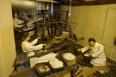 Jeju Folklor ve Doğa Tarihi Müzesi 'nde dönen ve dokuma tekniklerini gösteren iki manken gibi heddles ve warp iplikleriyle bir zemin tezgahı koza sepetlerinin yanında duruyor..