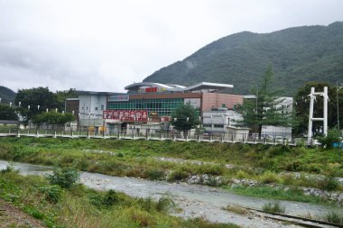 Mungyeong Geleneksel El Sanatları Müzesi, modern çatı ve dağ zeminine doğru uzanan yaya köprüsü ve sicim ışıklarıyla derenin karşısında duruyor..