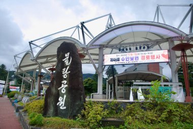 Kore harfleriyle oyulmuş uzun siyah bir taş tepe örtüsünün yanında duruyor ve renkli bir LED pankartı bir olayı duyuruyor, Suanbo Kaplıcaları 'nın giriş plazasını işaret ediyor..