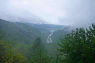 Ihwa Geçidi 'nin yakınındaki çam çerçeveli bir tepeden, vadi üzerinde puslu bir manzara açılıyor. Yol, katmanlı tepelerin altındaki dereyi takip ediyor ve yağmur üstüne bulutlar sürükleniyor..