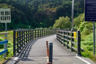 Uyarı ve bilgilendirme işaretleri Chuncheon 'da göl kıyısındaki bir rıhtımın girişinde duruyor, korkuluklar bisikletçileri ve sazlıkların üzerinden nehir kenarındaki ormana doğru yürüyenleri yönlendiriyor..