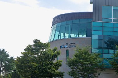 Robot Stüdyosu 'nun cam kubbesi Chuncheon' daki Animasyon Müzesi kompleksinin yanındaki ağaçların üzerinden yükseliyor. Geniş pencereler ve nehir kenarındaki parka bakan kıvrımlı kaplamalarla..