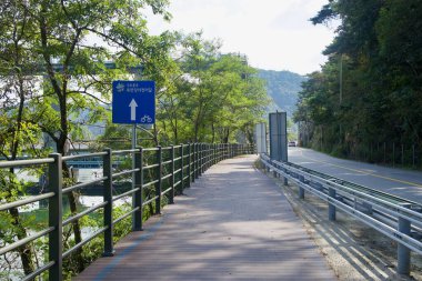 Bukhangang Bisiklet Yolu 'nun bir bölümü Gyeongchun Yolu' nun yanındaki korkuluk ve ağaçlar arasında yol alıyor. Mavi yol işareti Uiam Barajı 'nın kuzeyindeki nehir boyunca uzanan yolu gösteriyor..