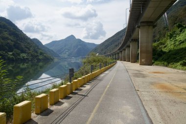 Sarı bloklar ve kablolu raylar, nehir yamaçlarını yansıtan ağaçlık yamaçlar ve uzak mesafede asılı bir köprü ile süpürme viyadüğün altındaki rıhtım yolu boyunca dizili..