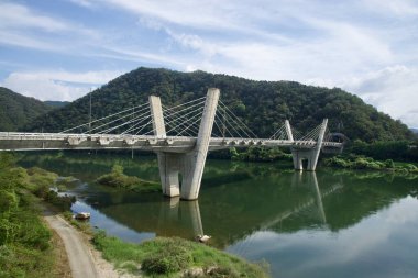 Yükseltilmiş bir bakış açısıyla, Gangchon Büyük Köprüsü, Bukhangang 'ı iki eğimli sütun ve yayılan kablolarla kaplar, ağaçlık tepelerin altındaki yavaş suya ve alçak bulut şeritlerine yansıtır..