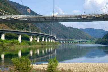 İkiz viyadükler, Gangchon Grand Köprüsü 'nün altındaki Kuzey Han Nehri kanyonunu takip ediyor. İskeleleri sakin sularda yansıyor. İki ziyaretçi ise bir çakıl barında duruyor..