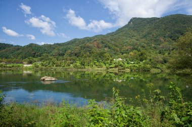 Kuzey Han Nehri 'nin geniş bir manzarası, ormanlık tepeler, yoğun nehir kenarı büyümesi ve parlak gün ortası gökyüzü olan alçak bir kayayı gösteriyor..