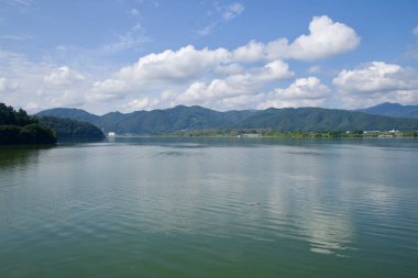 Uiam Gölü, mavi gökyüzünün altında batıya doğru uzanır. Nazikçe dalgalanan yüzeyi, geçen bulutları ve Chuncheon 'un etrafındaki tepeleri yansıtır..
