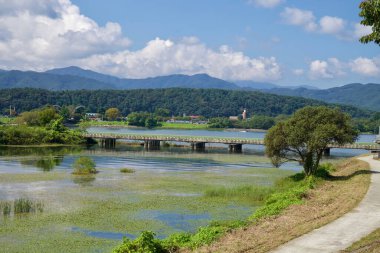 Alçak beton bir köprü, sualtı bitkileriyle dolu bir durgun suları geçerek ağaçlık tepelerin altındaki nehir kıyılarını birbirine bağlar ve Chuncheon 'da parlak bir öğleden sonra ince bir bulut yayılır..