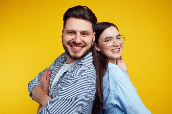 Couple stands backs and smiling against yellow background