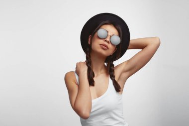 Beyaz gömlekli, güneş gözlüklü, şapkalı ve beyaz duvarda poz veren bir kız.