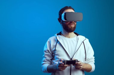 Heyecanlı sakallı genç adam, VR ekipmanlarıyla ayakta duruyor ve oynuyor.