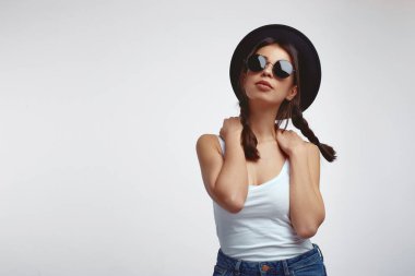 Beyaz gömlekli, güneş gözlüklü, beyaz duvarda poz veren şık bir genç kız.