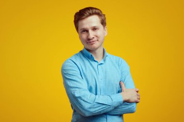 Mavi gömlekli adam ellerini turuncu arka planda çapraz yaparak mutlu poz veriyor.