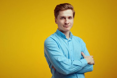 Mavi gömlekli adam ellerini turuncu arka planda çapraz yaparak mutlu poz veriyor.