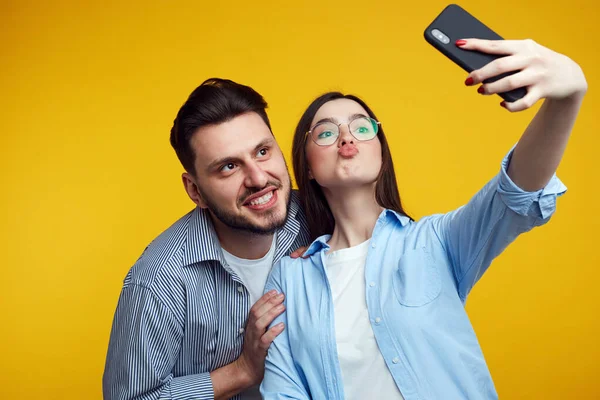 Komik çift somurtuyor, sarı duvarda akıllı telefondan selfie çekiyor.