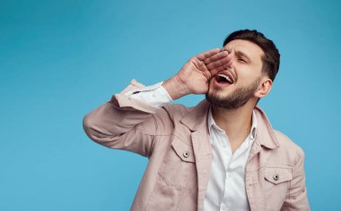 Mavi stüdyo arka planında eli ağzında bağıran genç yakışıklı bir adam.