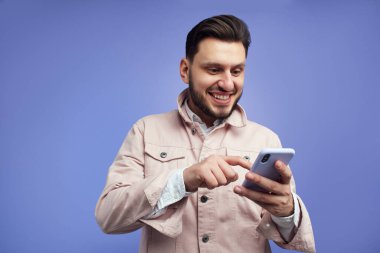 Akıllı telefon kullanan neşeli genç adam, mavi arka planda izole edilmiş.