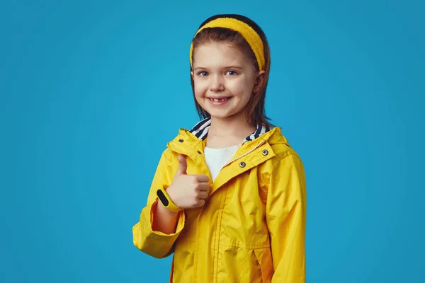 Linda chica con impermeable amarillo con capucha tiene buen humor, mostrando el pulgar hacia arriba — Foto de Stock