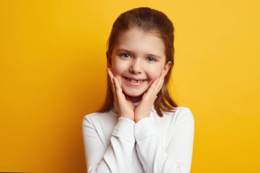 Genç ve tatlı kız ellerini yanaklarından ayırmıyor ve sarı duvara gülümsüyor.