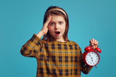 Şok olmuş, elinde çalar saat tutan kız mavi arka planda izole edilmiş.