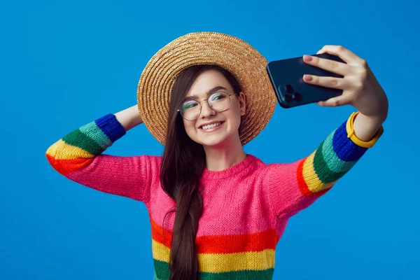 Gökkuşağı süveteri giyen bir kız cep telefonuyla selfie çekerken geniş bir gülümseme sergiliyor. — Stok fotoğraf