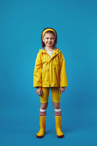 Full body girl in yellow raincoat and boots smiles while standing over blue