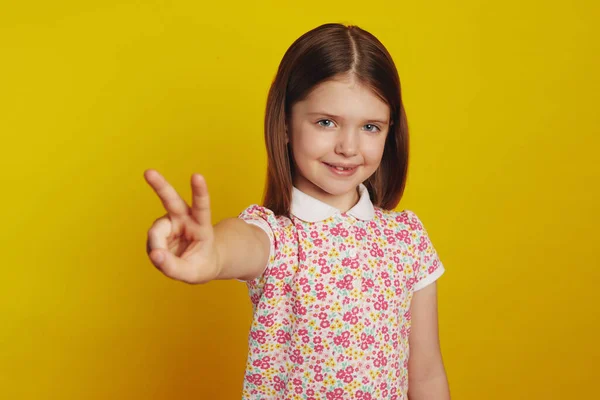 Niña sonriendo mientras muestra gesto de paz sobre fondo de estudio amarillo — Foto de Stock