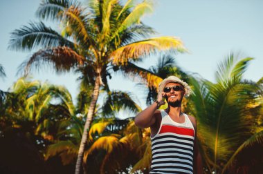 Akıllı telefondan konuşan sakallı genç adam palmiye ağaçlarının arka planında