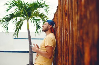 Elinde akıllı telefon tutan ve ahşap çitlere ve palmiye ağaçlarına karşı müzik dinleyen adam.