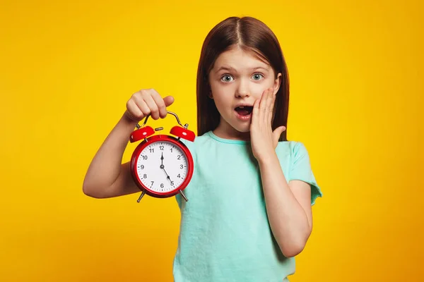 Sarı arka plan üzerinde alarmlı saat tutan şaşkın kız.