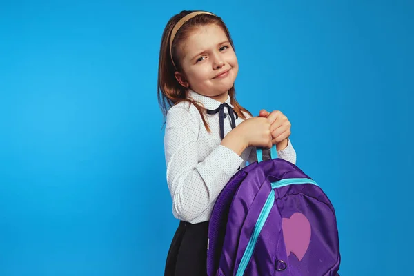 Симпатична школярка в уніформі тримає важкий рюкзак на синьому фоні — стокове фото