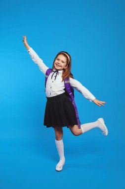 Üniformalı ve sırt çantalı kız ellerini mavi duvara kaldırıyor.