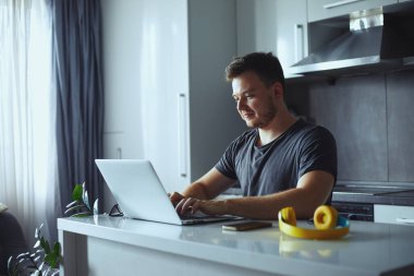 Gülümseyen serbest çalışan genç adam bilgisayarını kullanarak evde çalışıyor.