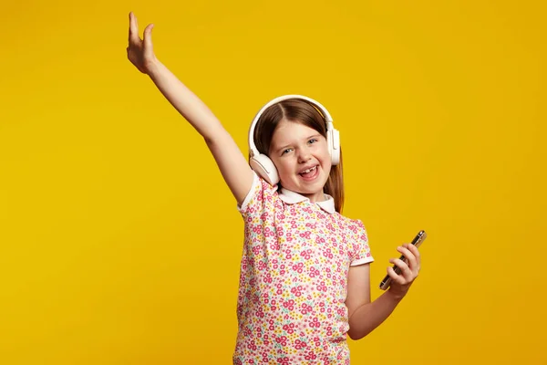 Menina com smartphone e fones de ouvido brancos levantando a mão, celebrando a vitória — Fotografia de Stock