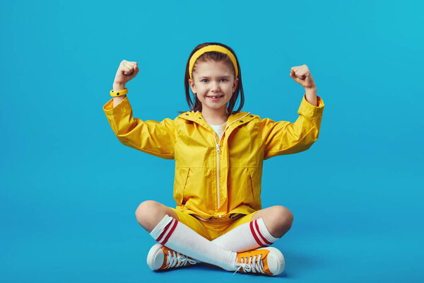 Girl raises hands and shows muscles dressed in yellow raincoat and headband
