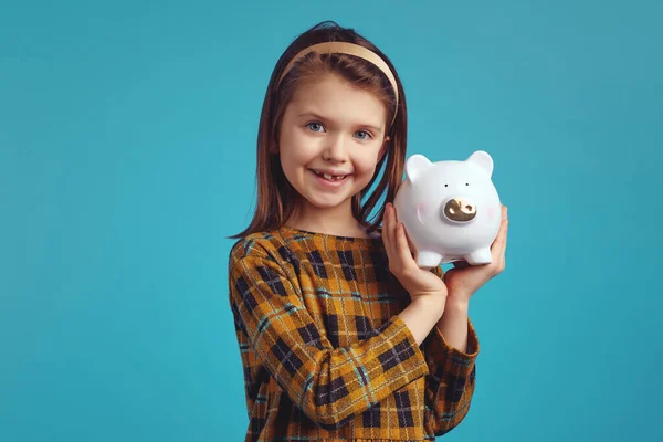 Küçük sevimli kız elinde domuz şeklinde para kutusu tutuyor. Mutlu görünüyor.